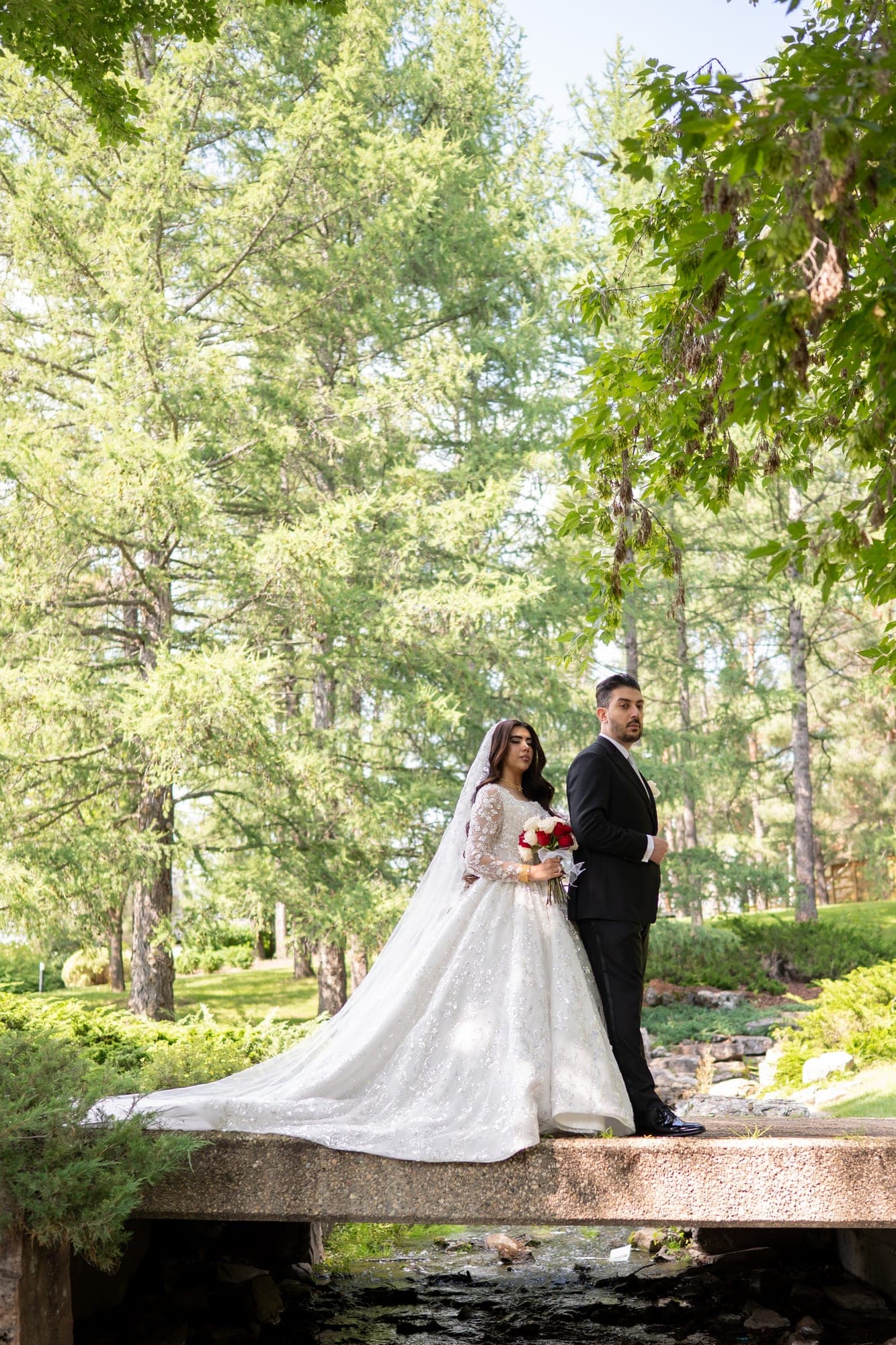 A bride and groom standing together on a small bridge surrounded by tall green trees