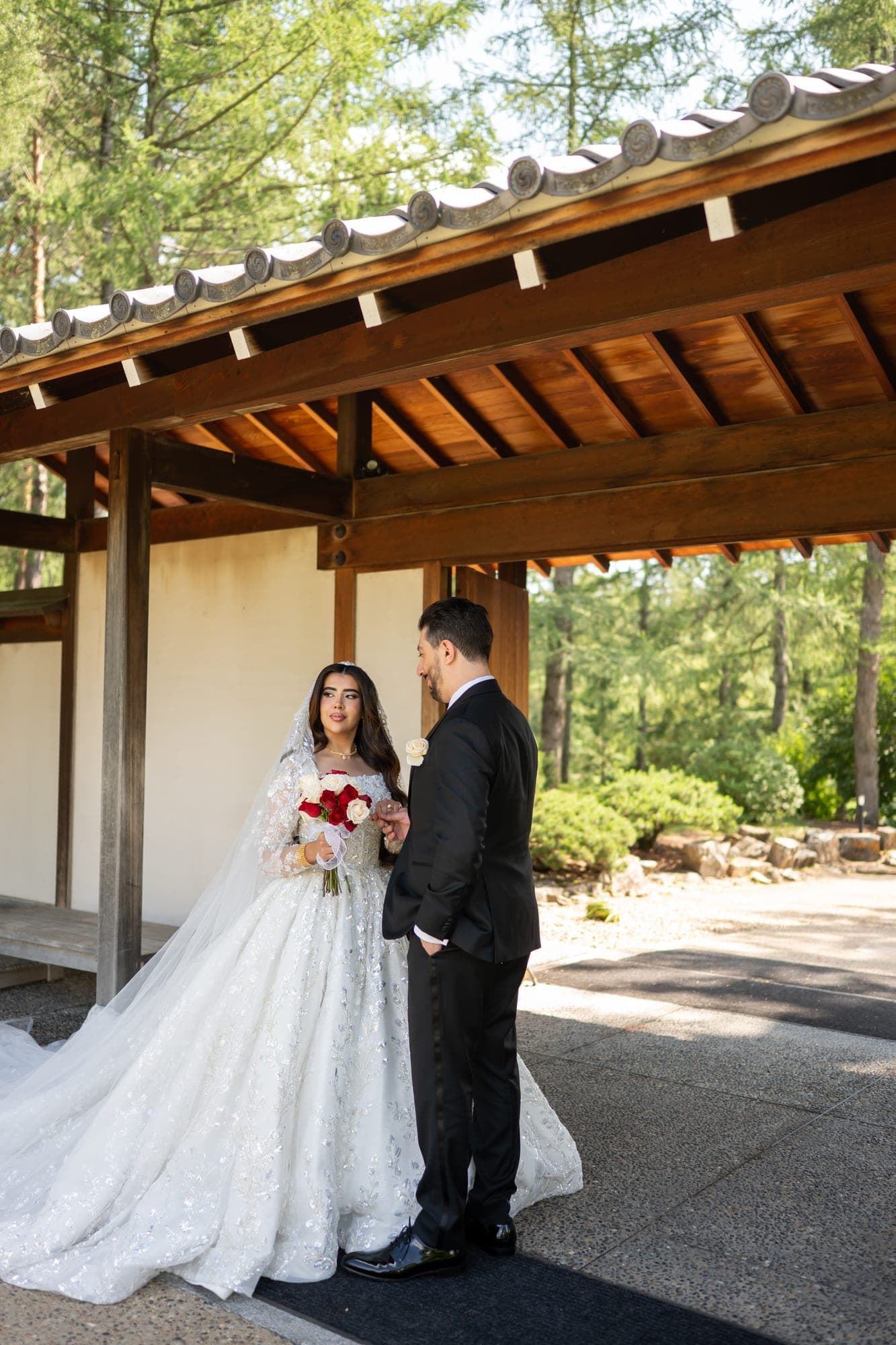 A bride holding red and white roses beside the groom beneath a timber garden pavilion