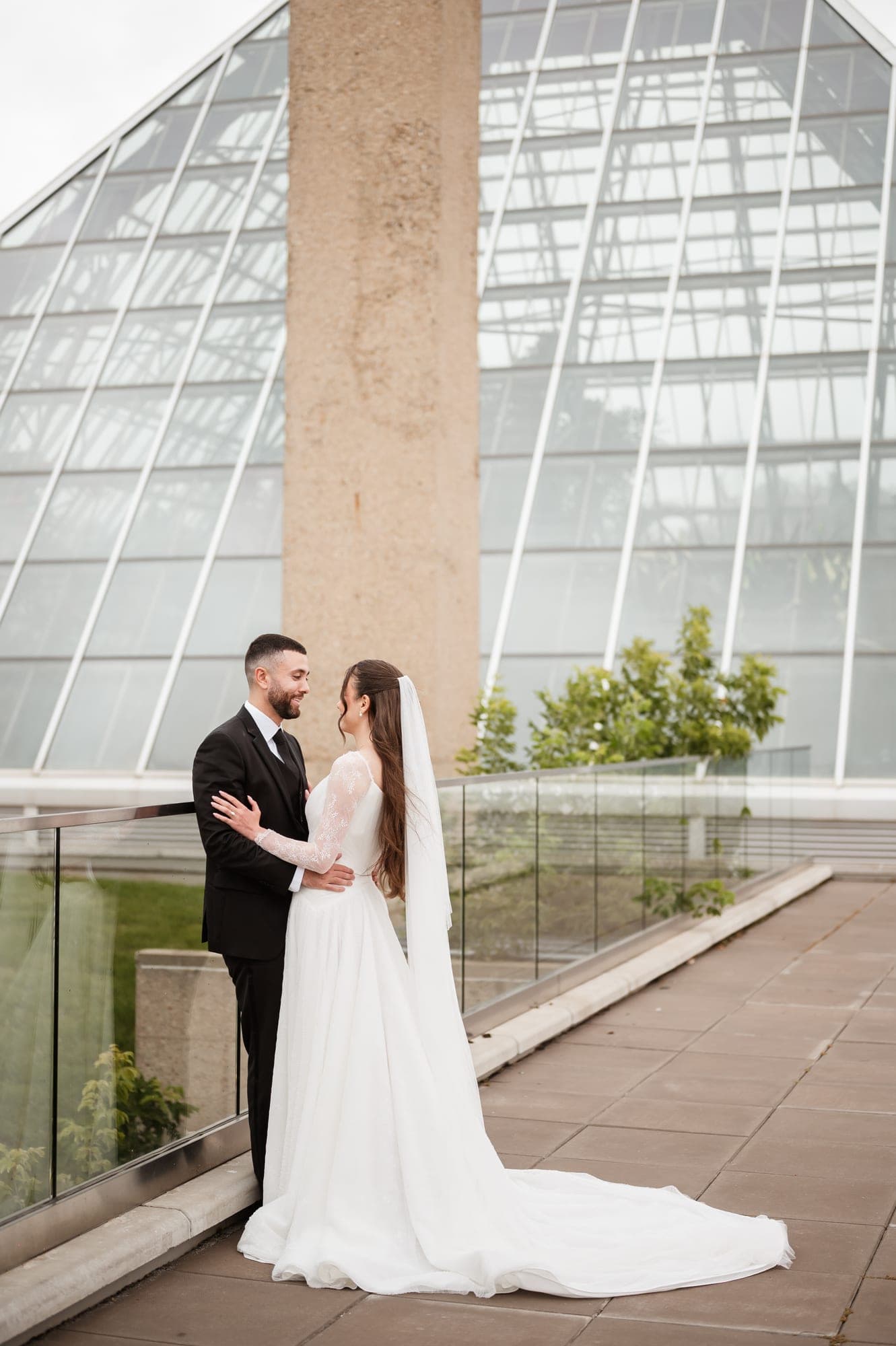 A newlywed couple standing together on a terrace outside a modern glass conservatory