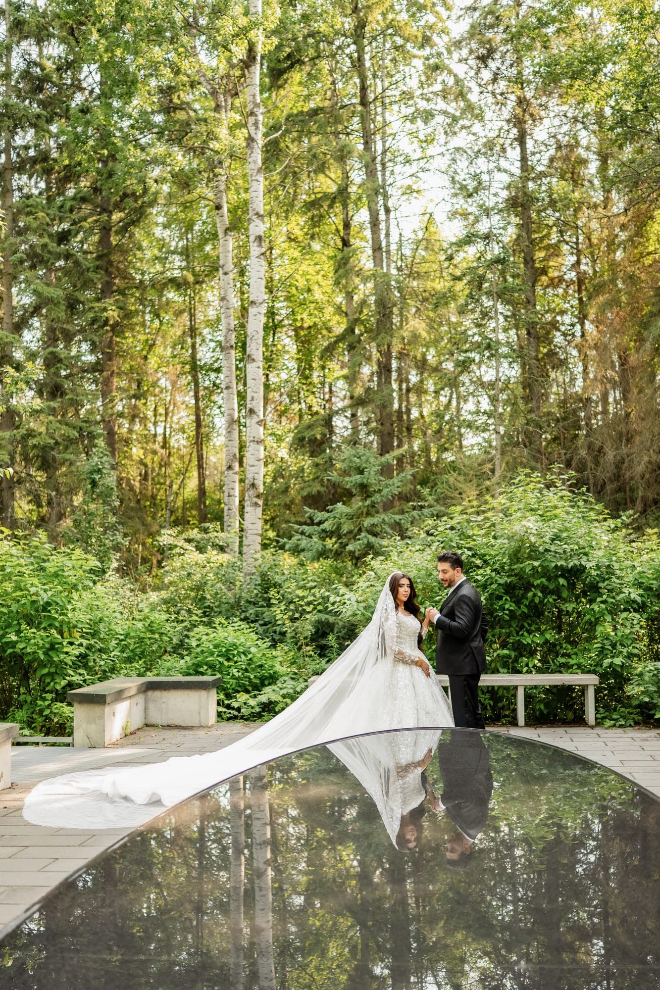 A bride and groom reflected in a dark garden pool beneath tall woodland trees