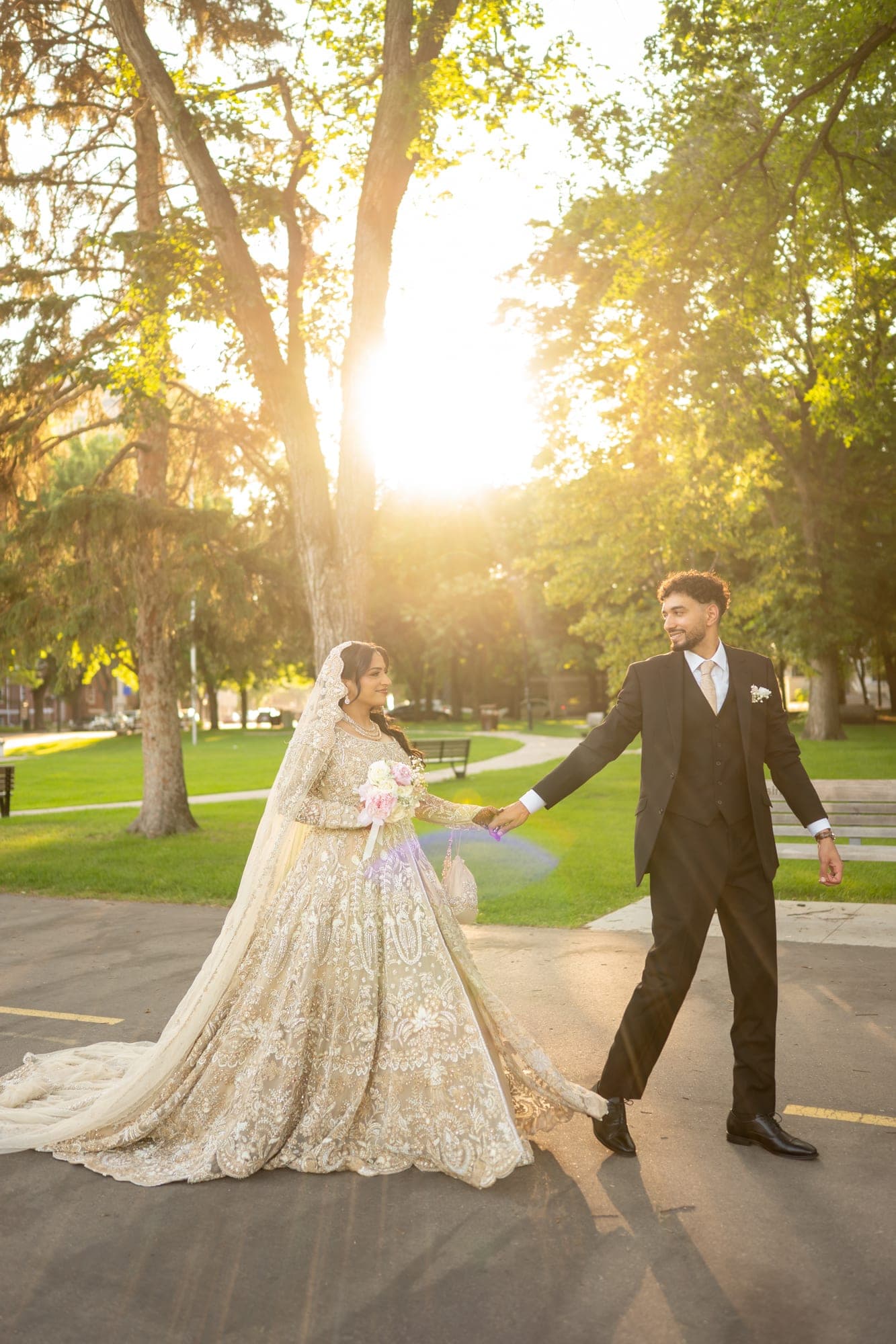 A newlywed couple holding hands in a park as warm evening sunlight filters through the trees
